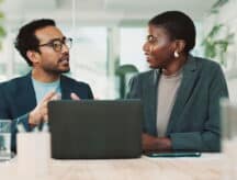 Man and woman having a discussion in an office environment, with a laptop and glass of water in front of them.