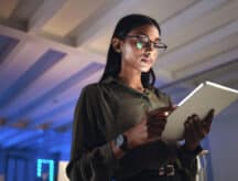 A woman in glasses standing and holding a tablet in her hand, looking serious. There is a blue glow behind her.