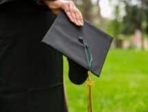 A hand of a woman in a graduation gown holding a morarboard in a park after graduation.