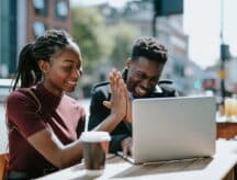 A couple sitting outside at a table, which has a laptop and a coffee cup on it, giving each other a high five.