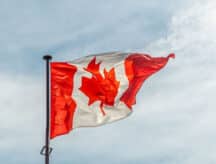 Canadian flag waving in the wind against a blue sky with some cloud coverage.