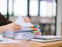 A person sitting at a wooden table and sorting through a stack of papers that have been grouped with coloured paper clips.