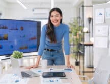 Woman leaning on desk, looking confident, surrounded by papers and electronics.