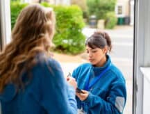 A woman standing in a door way talking to another woman who is writing on a tablet.
