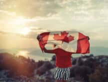 Teenage girl facing away from the camera, holding up the Canadian flag above her shoulders, at sunset.