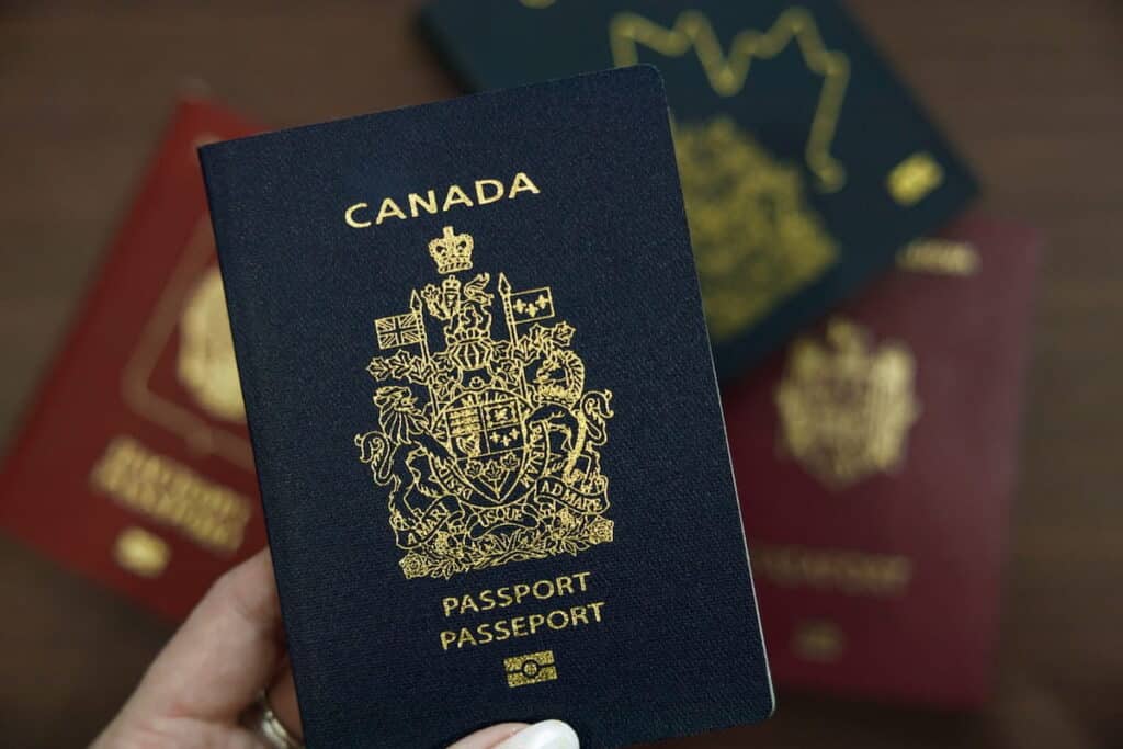 A hand holds a Canadian passport to the camera, as other passports make up the background.