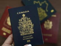 A hand holds a Canadian passport to the camera, as other passports make up the background.