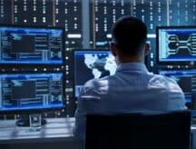 A man sitting in front of five computer screens which display varying bits of information.