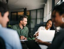 Group of workers laughing together in an office environment, pens and laptops before them.