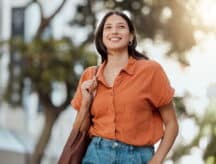 A shot of a happy woman in an orange shirt standing with one hand on the brown purse on her shoulder and another in the pockets of her jeans.