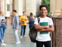 Man with a backpack over one shoulder and books in his hand, standing and smiling into the camera.