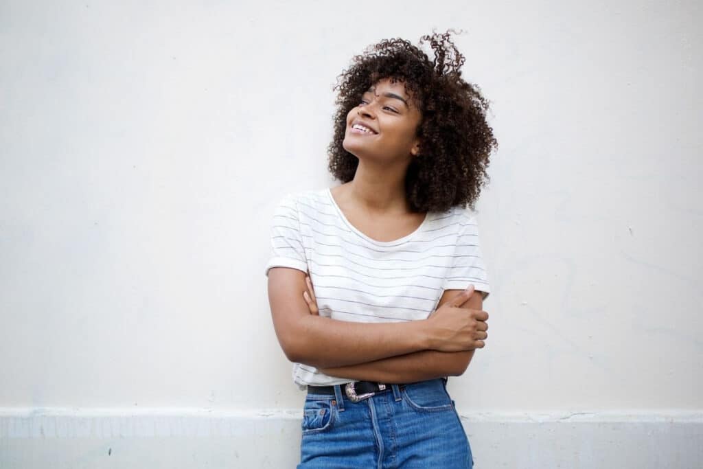 Woman leaning against a white wall, smiling and looking up to her right.