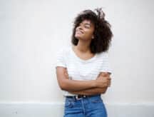 Woman leaning against a white wall, smiling and looking up to her right.