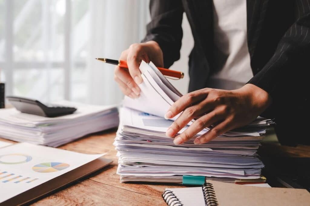 A closeup shot of someone's hands sorting through a stack of papers, with a red pen in their right hand.