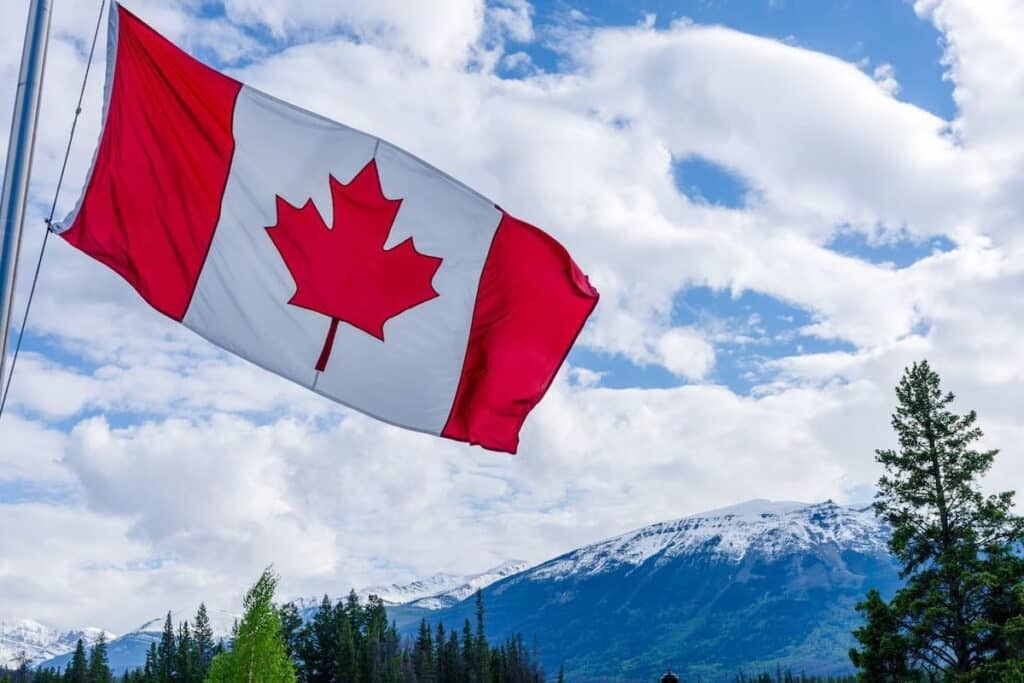 Closeup of Canadian flag waving in the wind, against a backdrop of snow-capped mountains and coniferous trees. Cloudy day.