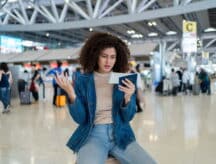 Woman in airport sitting on her luggage, looking at her passport and boarding pass, and throwing up a hand in confusion.