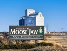 A "Welcome to Moose Jaw - The Friendly City" sign shot in front of a grain elevator on a day with a clear sky.