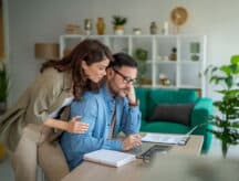 A couple looking at a laptop together; the man is sitting, while the woman stands behind him and holds his shoulders—faces show distress.