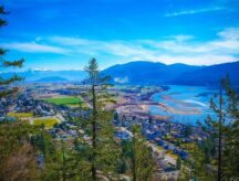 An aerial shot of greenery and homes against the Fraser Riser in British Columbia, with mountains visible in the background.