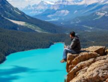 A man sits on the cliff-edge of a mountain looking down at Lake Morraine in Banff national park.