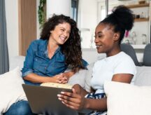 Two female friends sitting on the couch, very happy, while one shows the other something on a laptop.