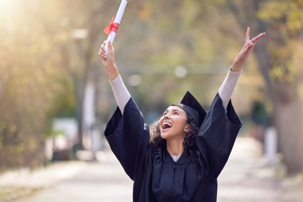 Young woman that just graduated jumping for joy in her graduation cap and gown, holding her degree.