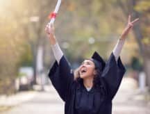 Young woman that just graduated jumping for joy in her graduation cap and gown, holding her degree.