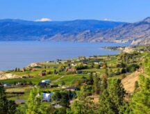 Naramata, a community within the Regional District of Okanagan–Similkameen, in British Columbia, Canada. Houses, greenery, and mountains are visible; clear blue skies.