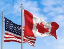 A closeup shot of the American flag and Canadian flag waving in the wind against a blue sky.