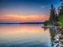A calm lake in Manitoba, with trees to the right, and colourful skies.
