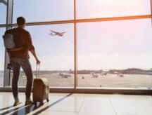 A man standing in an airport, looking out the window at a passing aircraft.