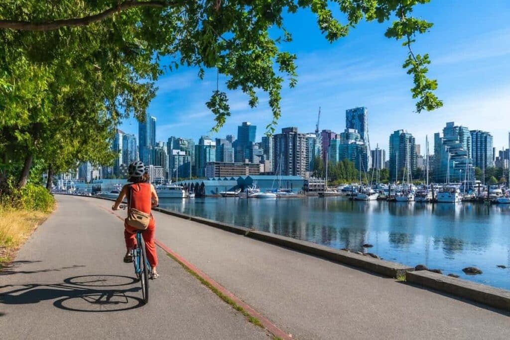 A person riding a bicycle along the Stanley Park Seawall, with the downtown Vancouver skyline in the background.