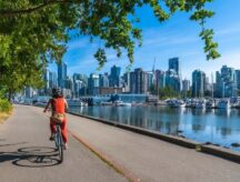 A person riding a bicycle along the Stanley Park Seawall, with the downtown Vancouver skyline in the background.