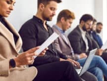 People lined up against a wall, waiting, while reading papers or their laptop.