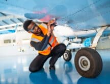 Aircraft maintenance engineer wearing safety glasses and a high-visibility vest inspects the wing of a small airplane inside a hangar.