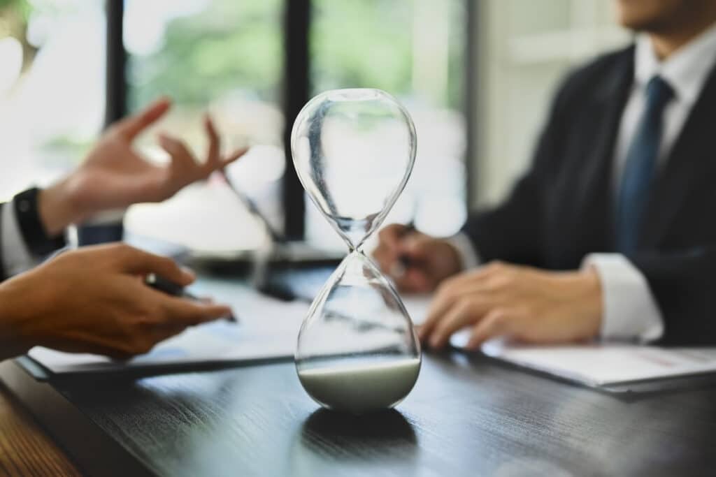 Closeup of an hourglass on a table as two businesspeople converse in the background.