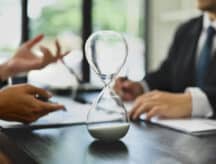 Closeup of an hourglass on a table as two businesspeople converse in the background.