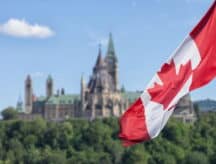 Canadian flag waving with Parliament Buildings hill and Library in the background.