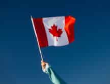 A hand holding up a Canadian flag against a blue sky.
