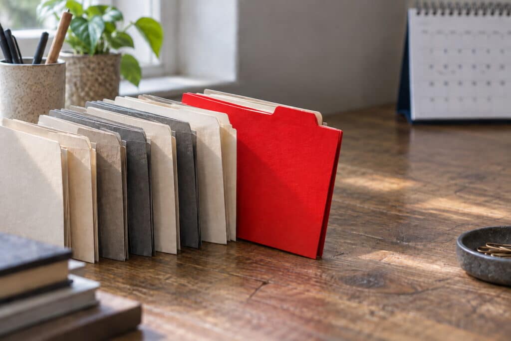 A row of files sits atop a busy desk with one red file sticking out.