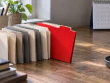 A row of files sits atop a busy desk with one red file sticking out.