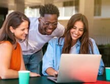 Three friends, smiling, huddled around a laptop.