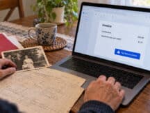 A person at a kitchen table pays an online invoice on a laptop, beside an old family photo and a handwritten birth register.